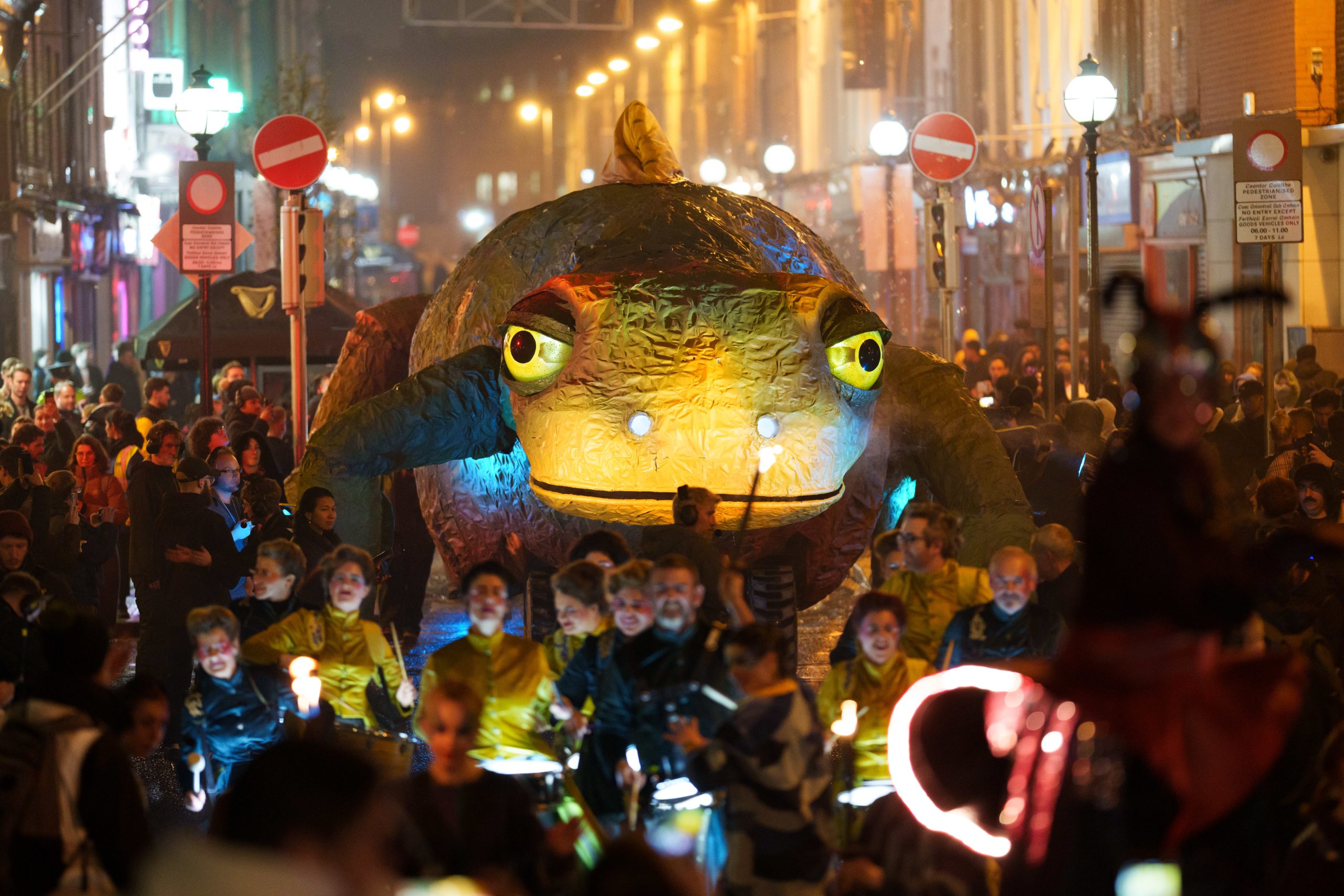 Large glowing Newt float in a nighttime street parade with performers and crowd.