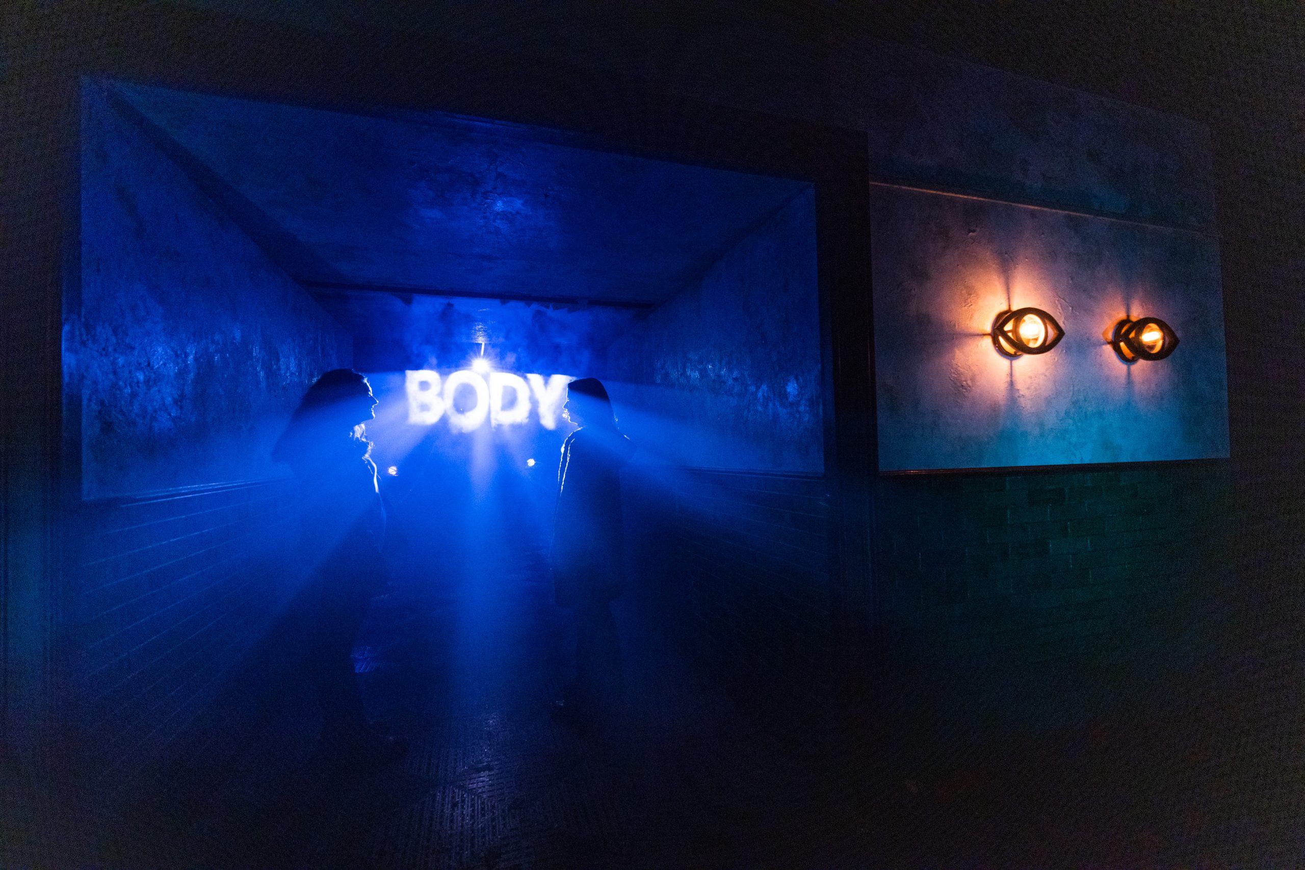 Two people facing a glowing "BODY" sign in a dark, misty room.