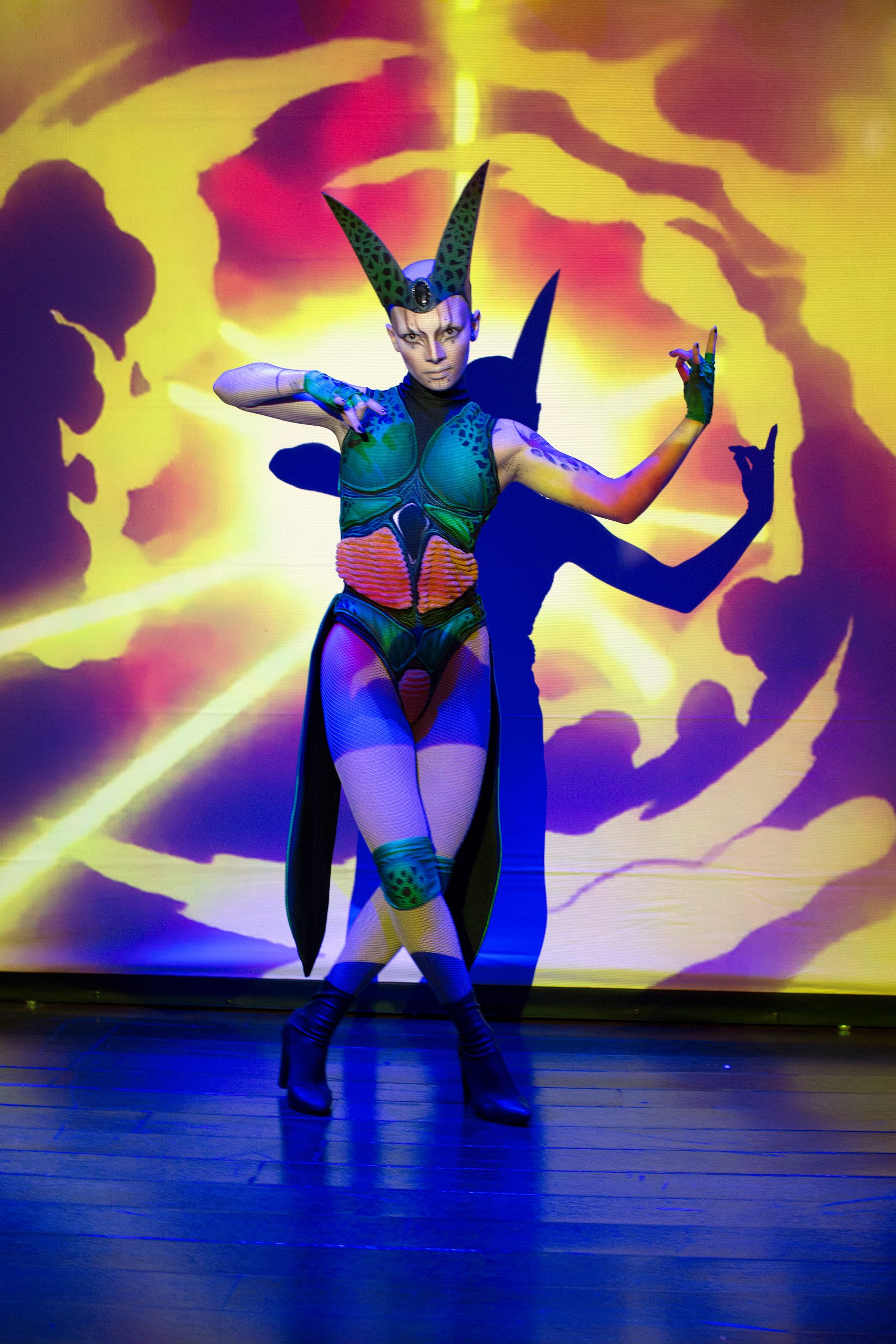 Performer in a colourful costume and horned headpiece posing in front of fiery background projection.