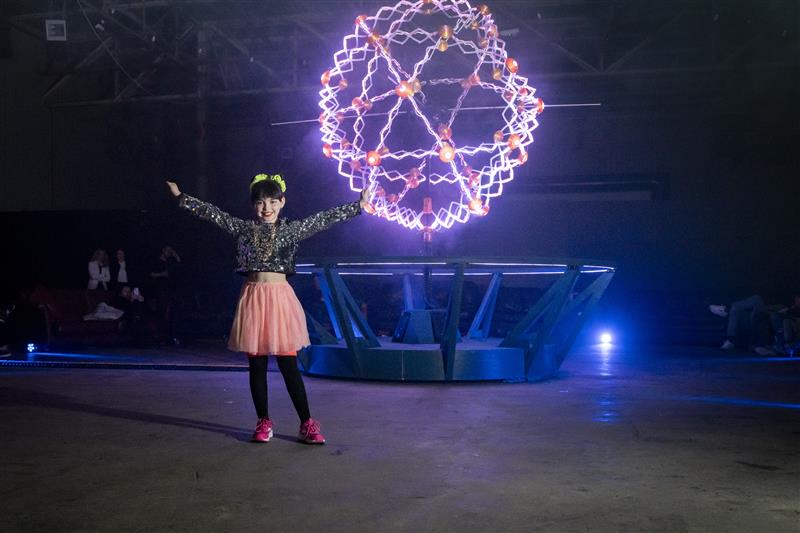 Young girl stands in front of illuminated geometric sphere sculpture with red and white lights in a dark exhibit space.