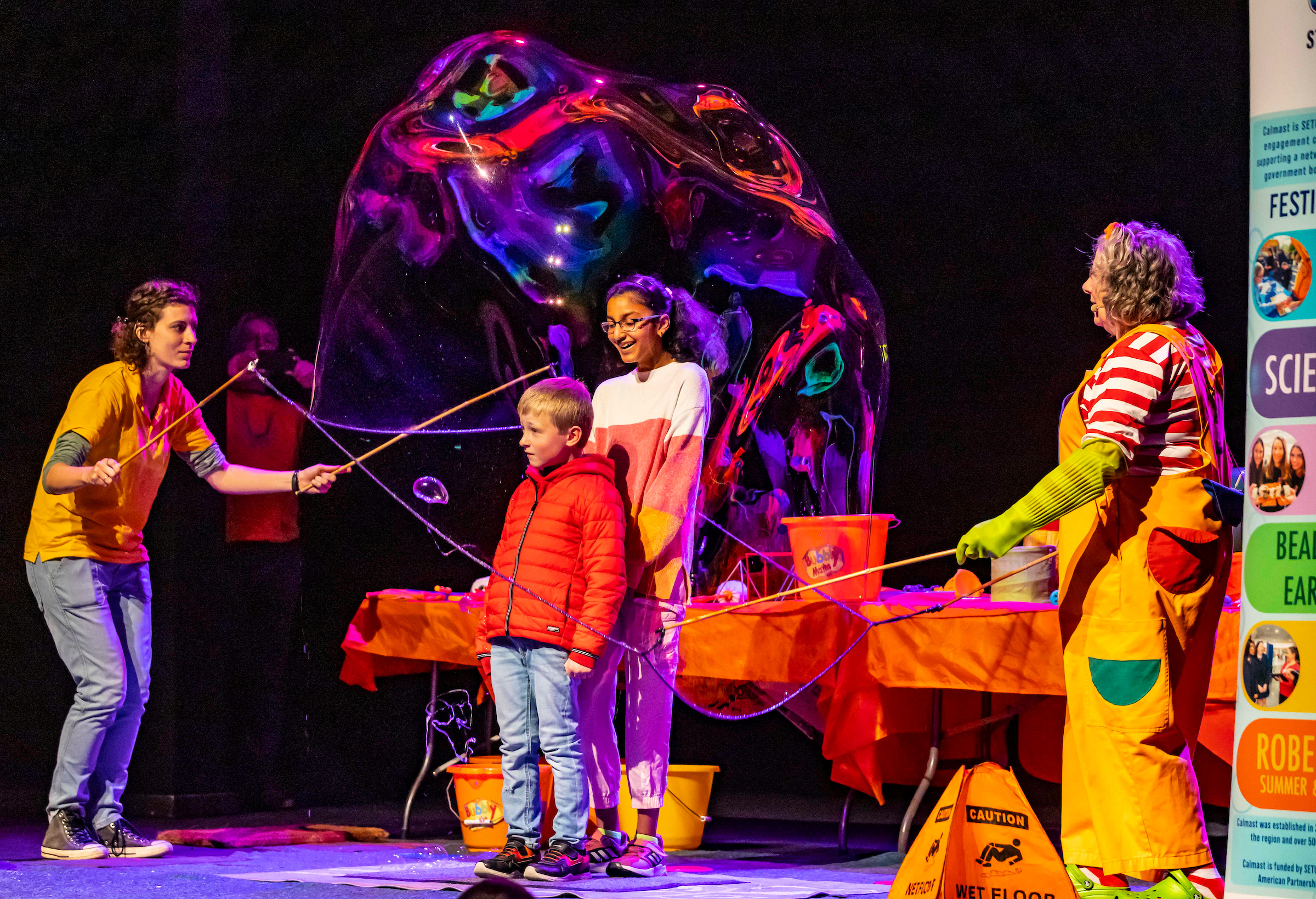 Two adults performing a bubble making activity with two children inside a large bubble surrounded by colourful lighting.