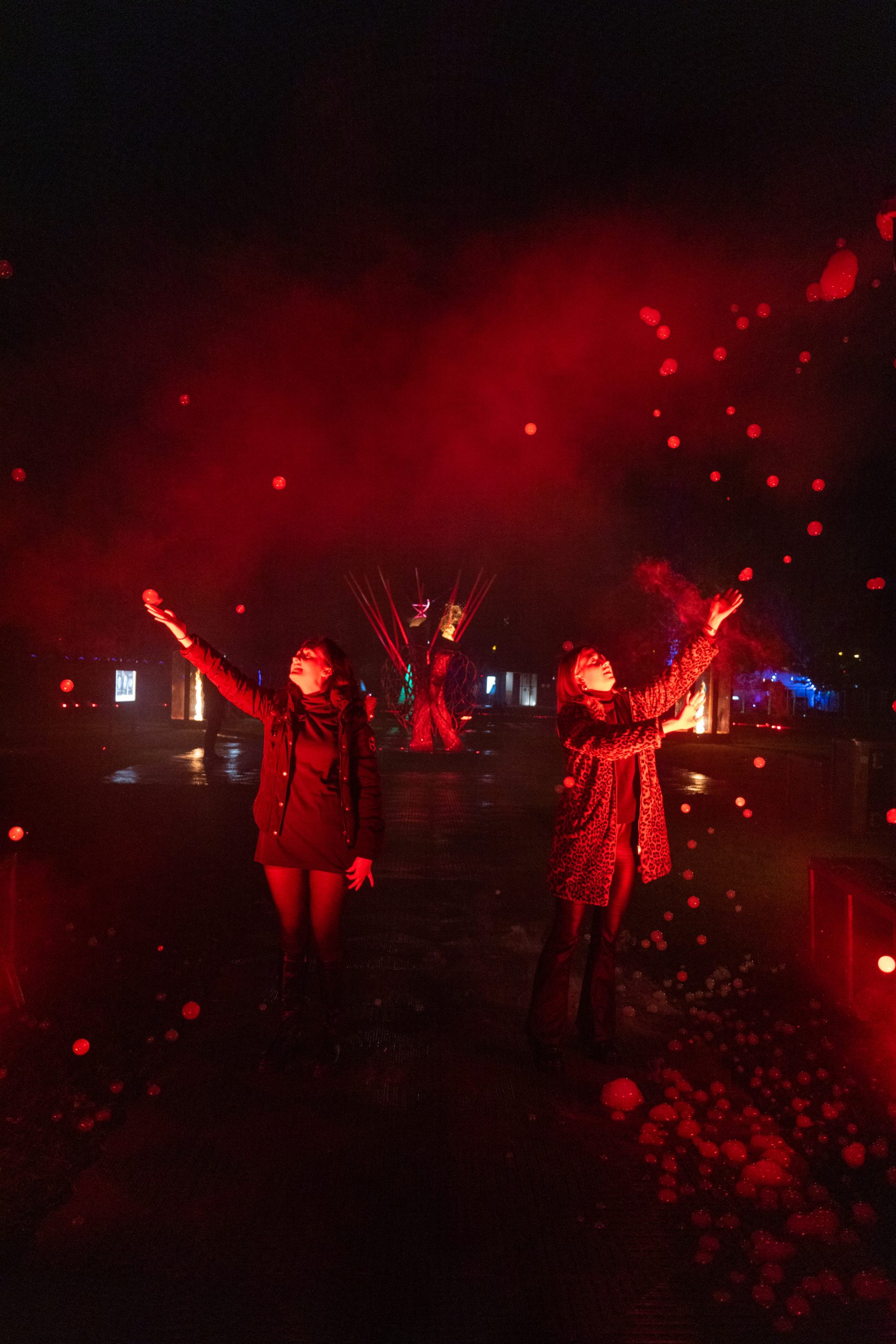 Two people standing outdoors at night, illuminated by red lighting and surrounded by floating bubbles. Behind them are colorful lights and an artistic installation with converging beams, reflected on the wet ground