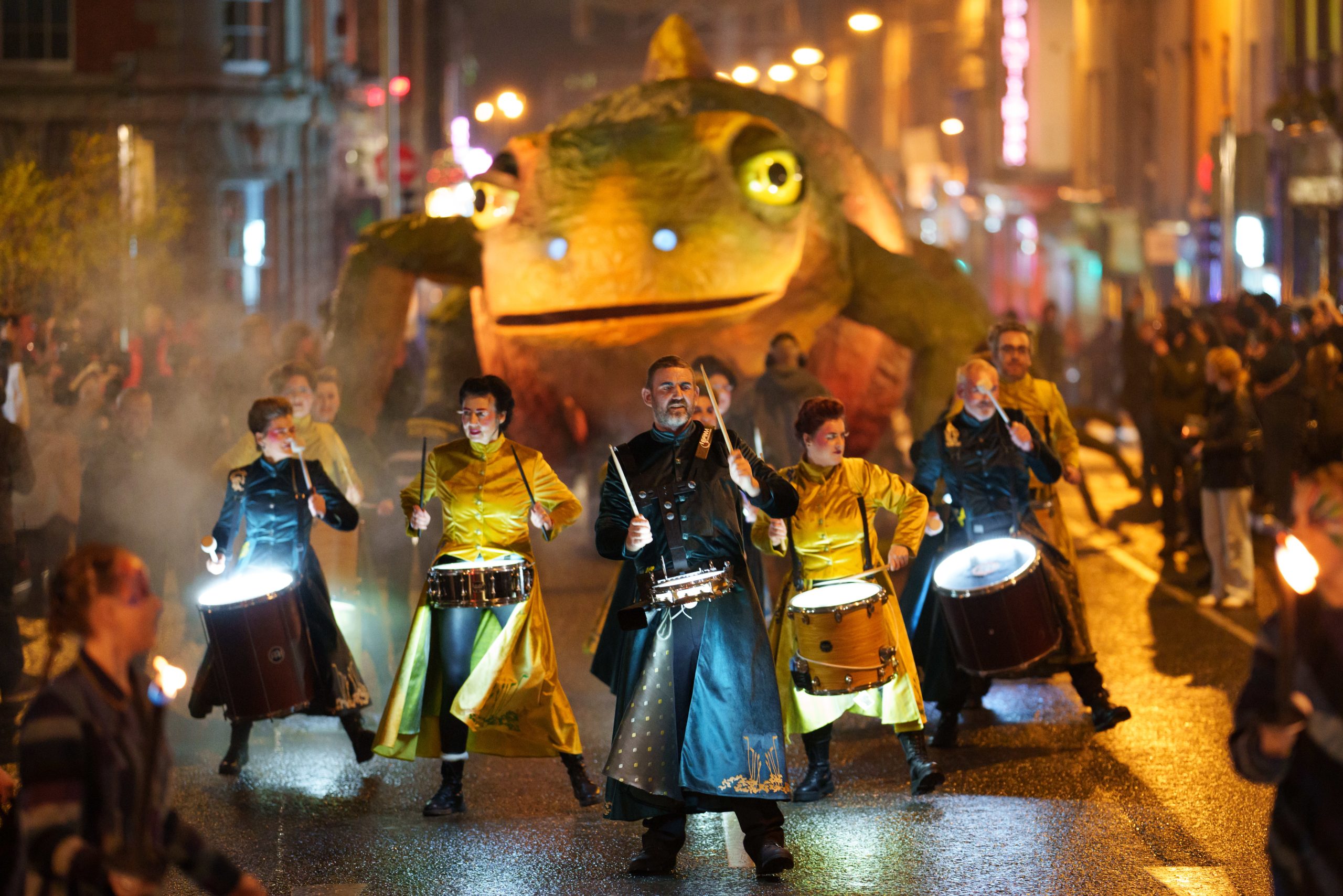 Drummers in colorful costumes perform in a night parade with a glowing newt float, watched spectators in the street.