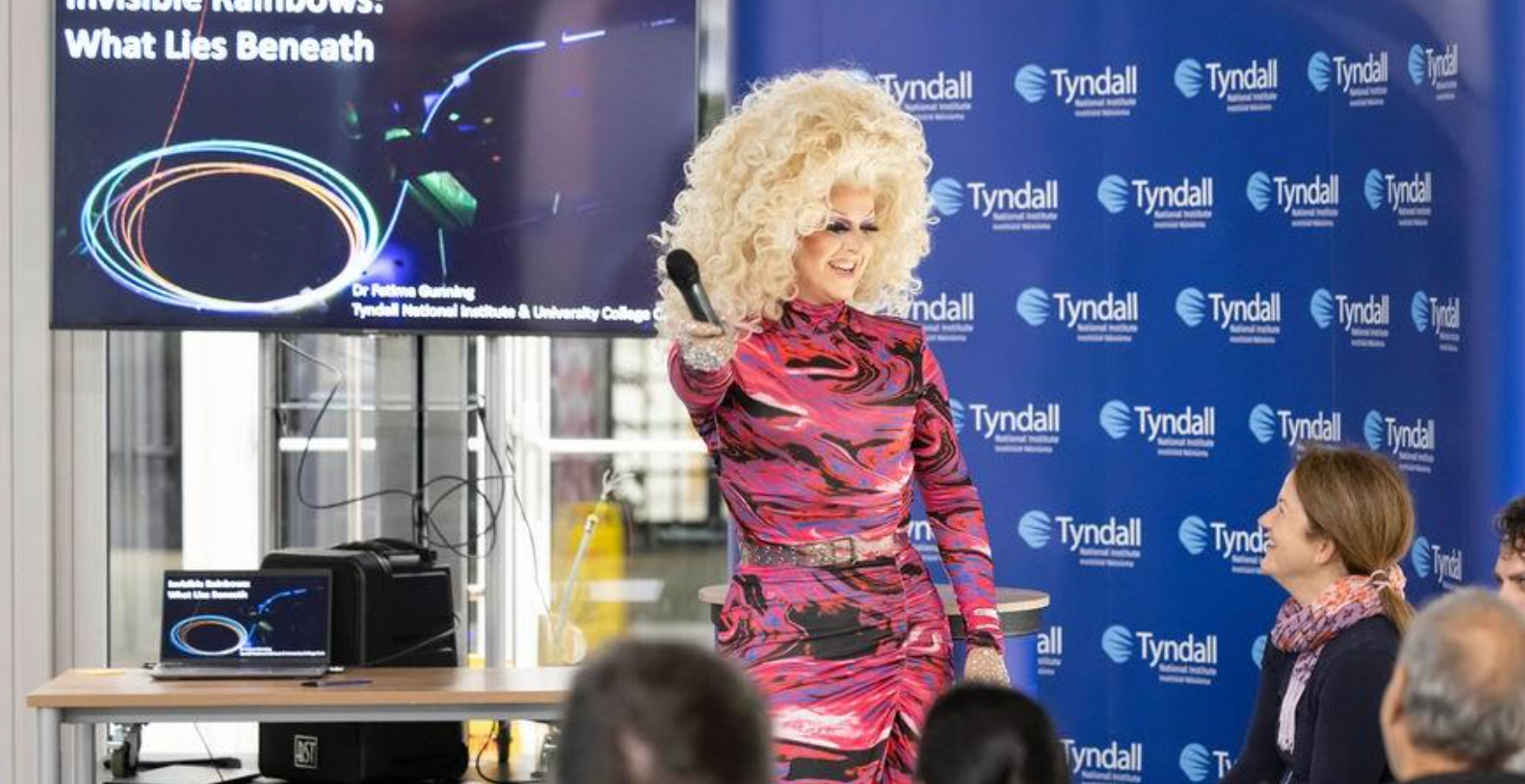 Speaker with curly blonde hair in a pink and black outfit, presenting a science event on stage with a Tyndall Institute banner behind