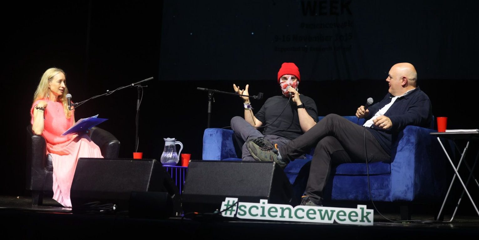 Three people sitting and talking on stage with handheld microphones in a tent.