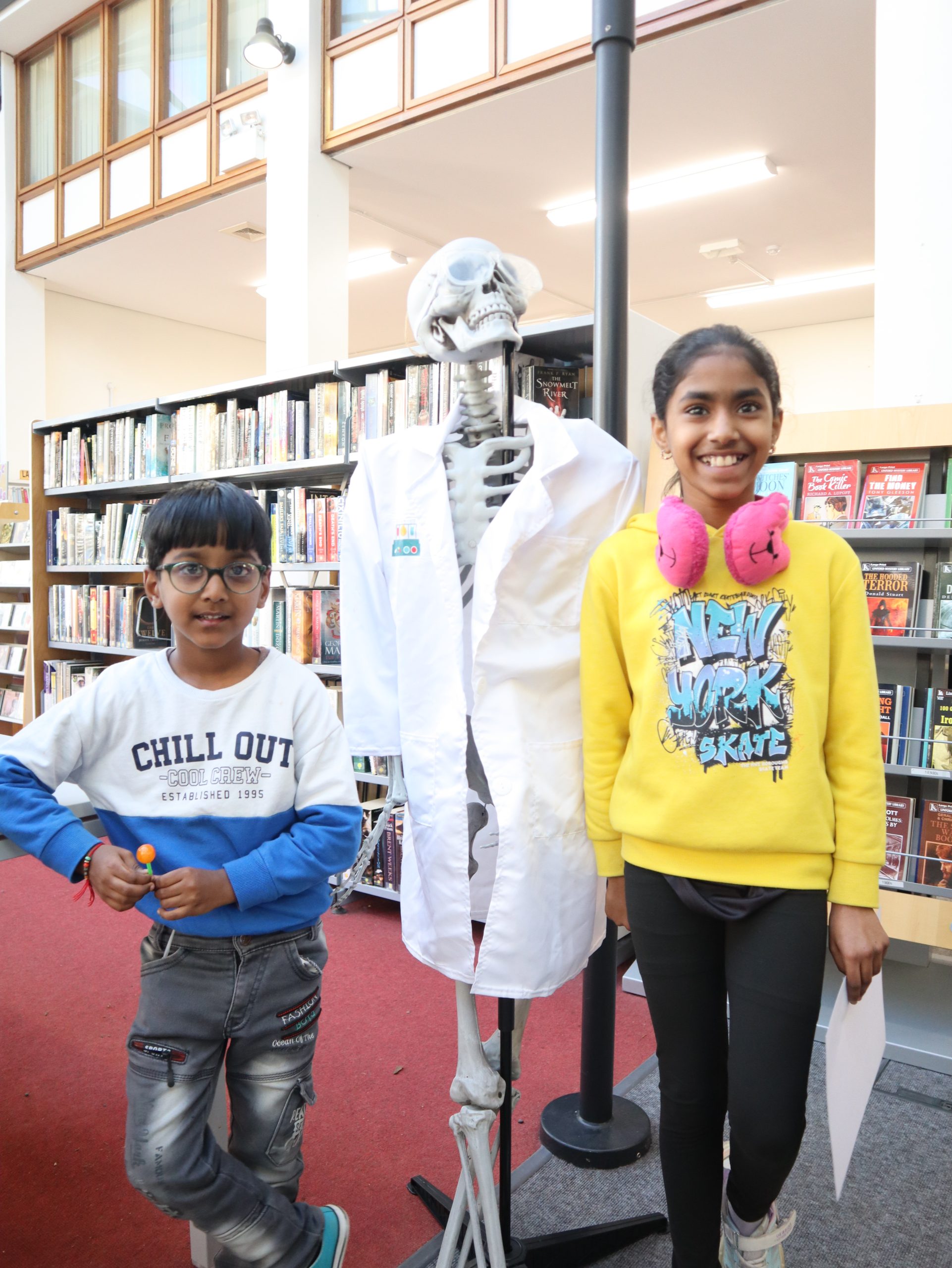 Two children stand beside a skeleton in a lab coat in a library. One wears a 'CHILL OUT' shirt and silver trousers; the other has a yellow sweatshirt and pink headphones.