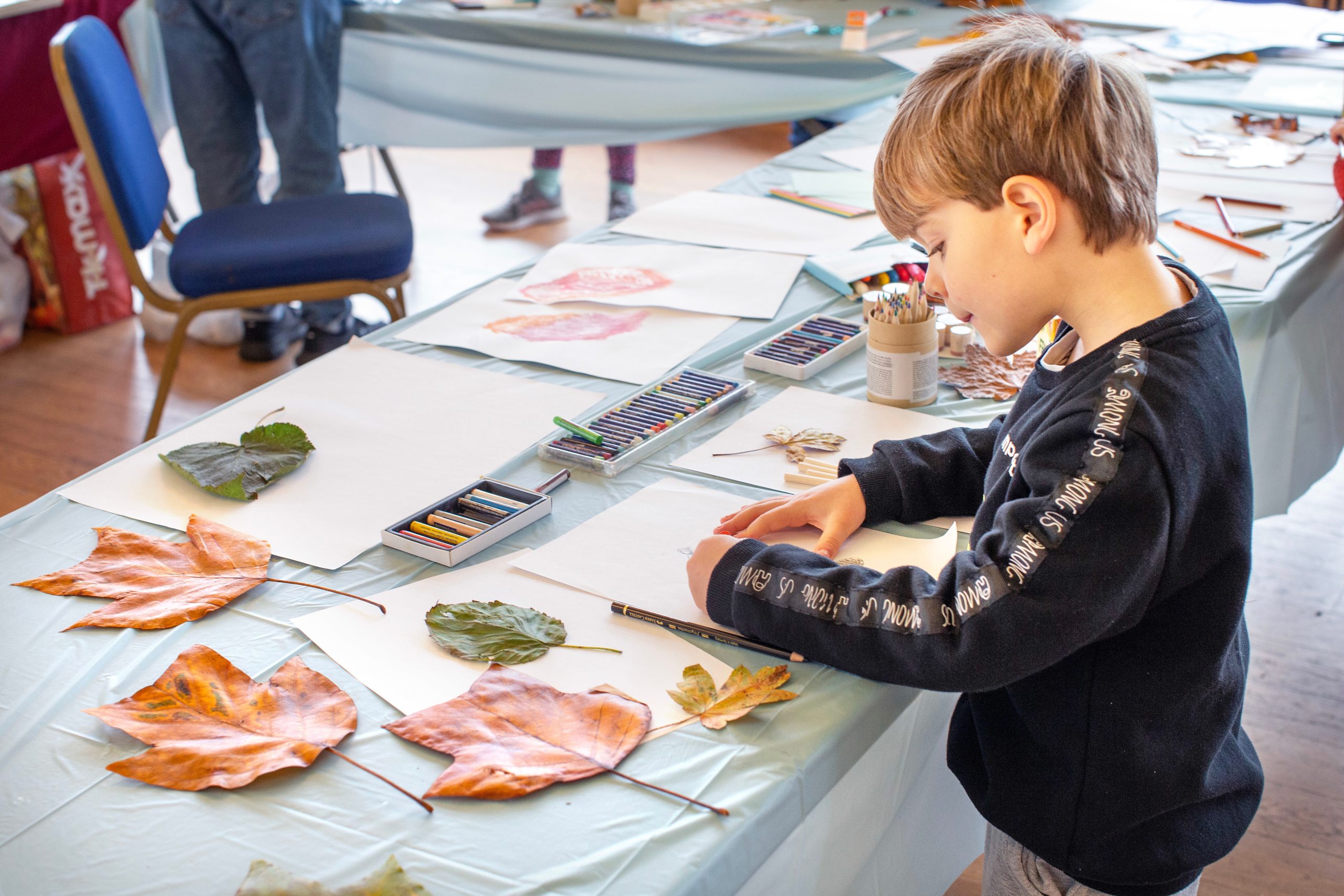 Child doing leaf art at a table with colored pencils and paper.