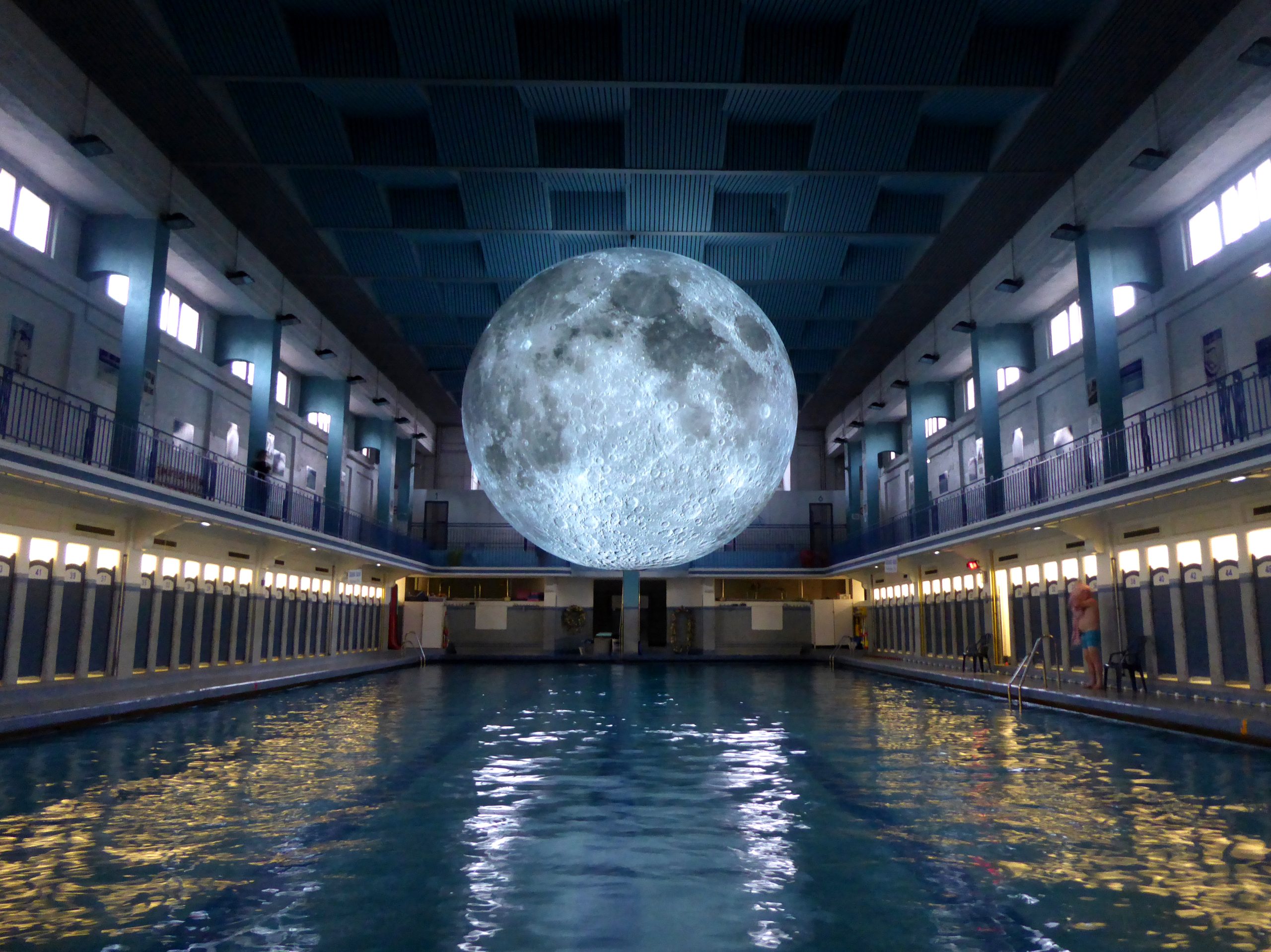An illuminated moon sculpture hangs above an indoor swimming pool, reflecting on the water below in a brightly lit space with a high blue-paneled ceiling