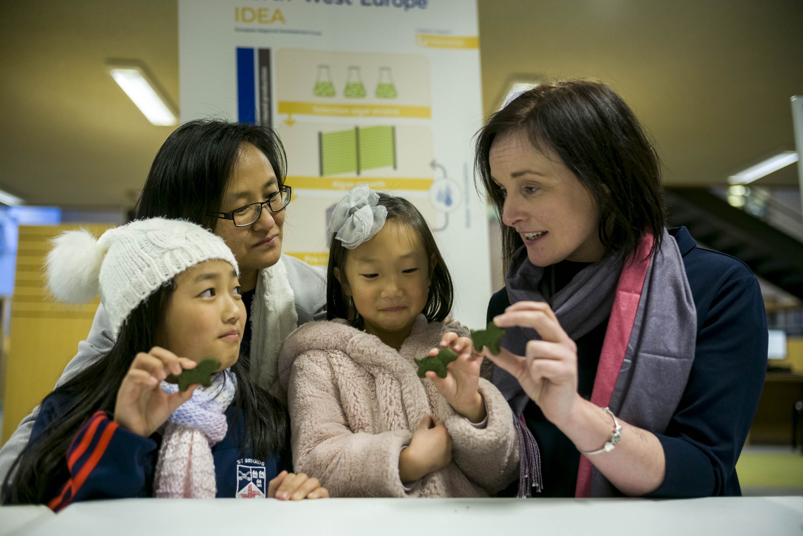 Two adults and two children examining small objects together indoors, with a poster in the background.