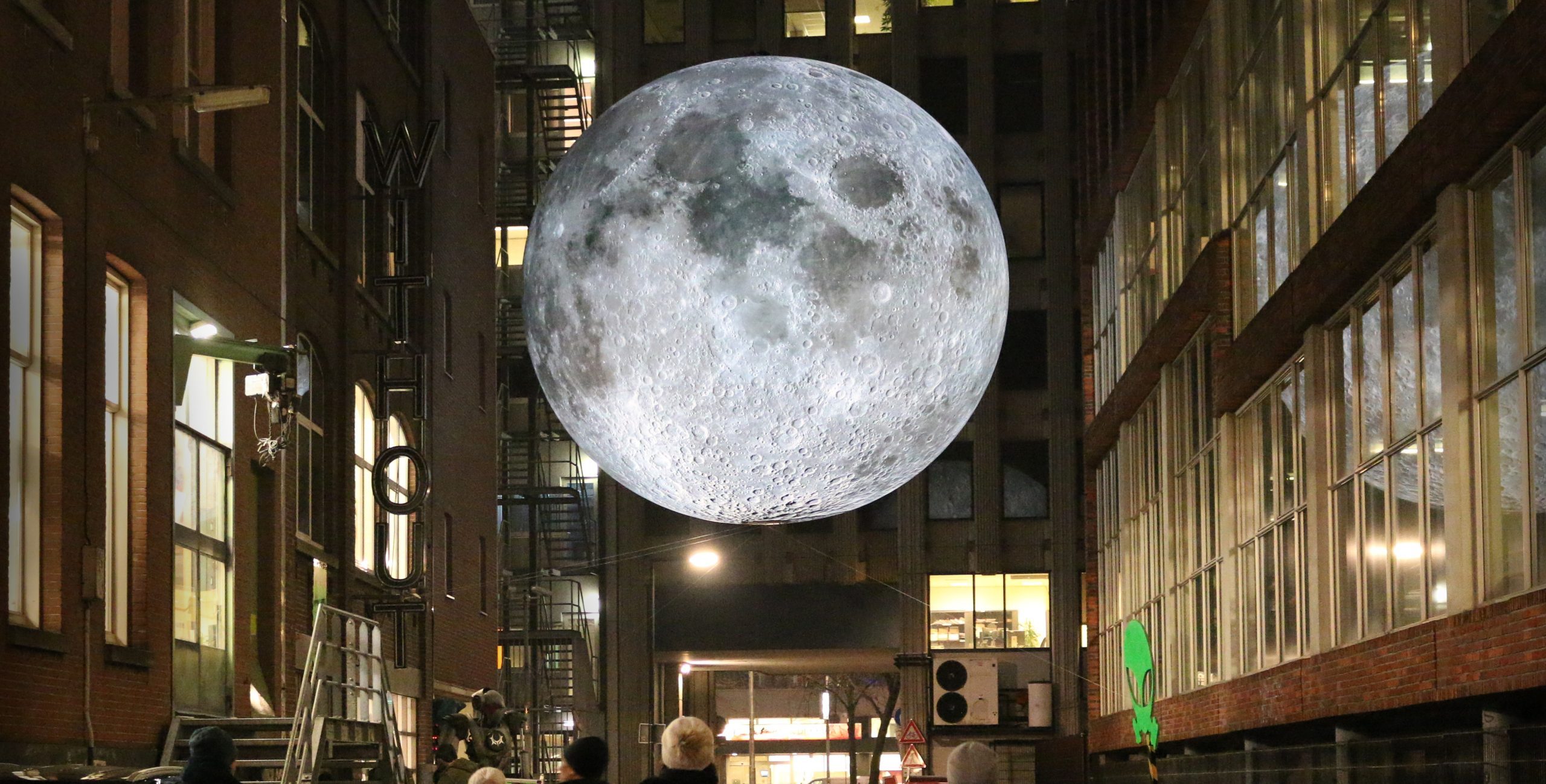 Nighttime urban scene with people walking beneath a large illuminated moon installation suspended between tall buildings.