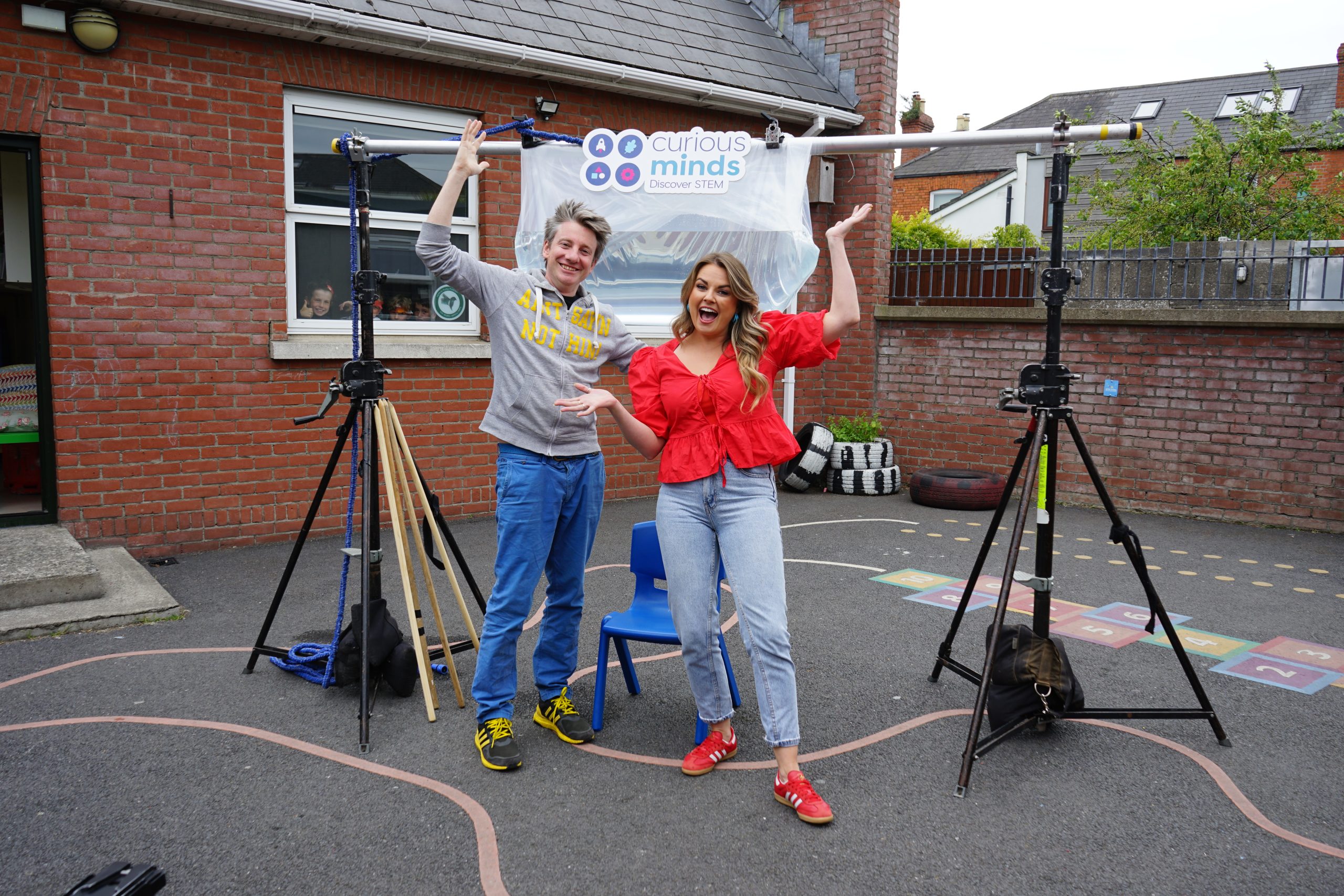 Two people standing outside a brick building, holding a "Curious Minds" banner, with hopscotch markings and tripods nearby.
