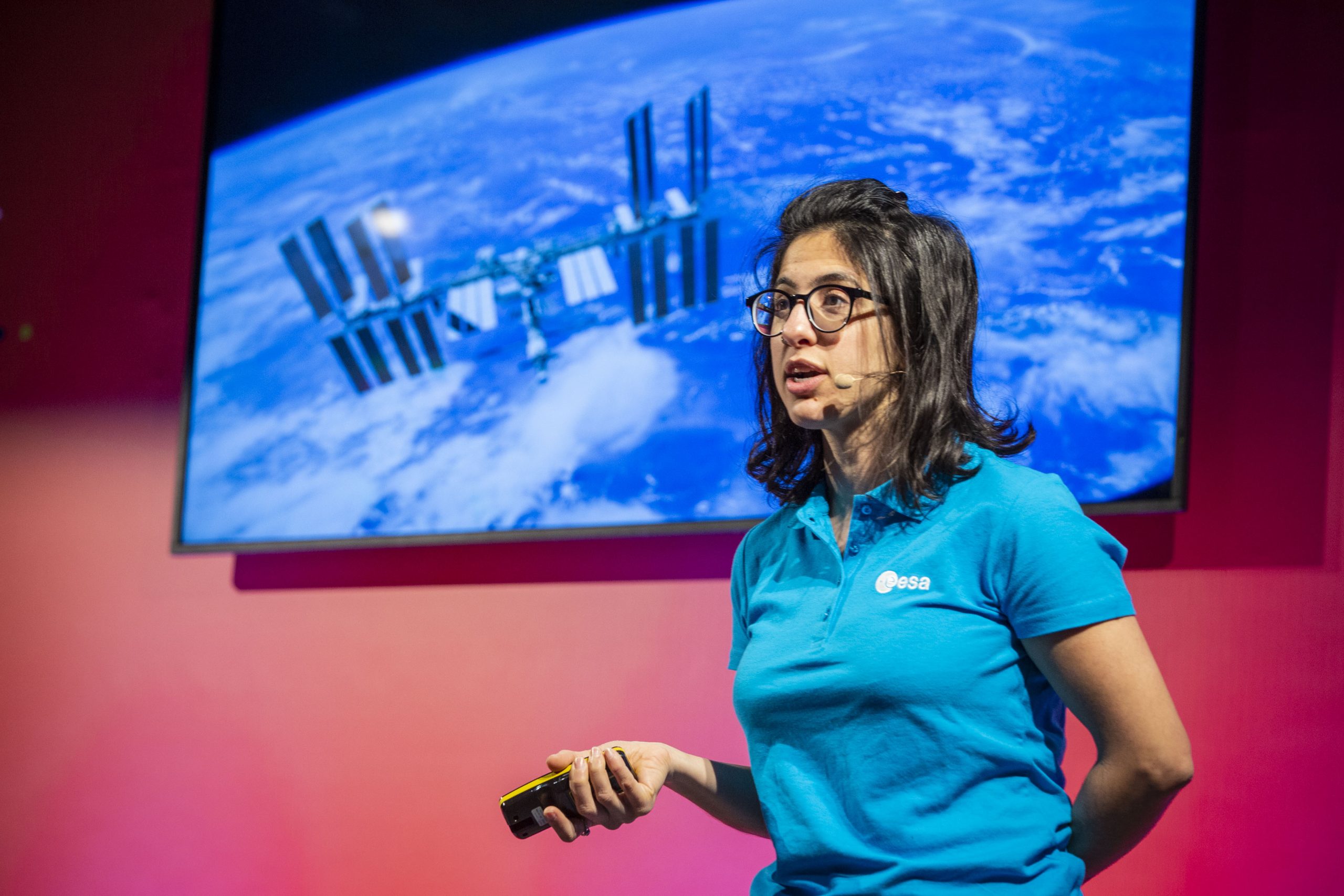 Person in ESA shirt presenting with a device in hand, standing in front of a screen showing the International Space Station above Earth.