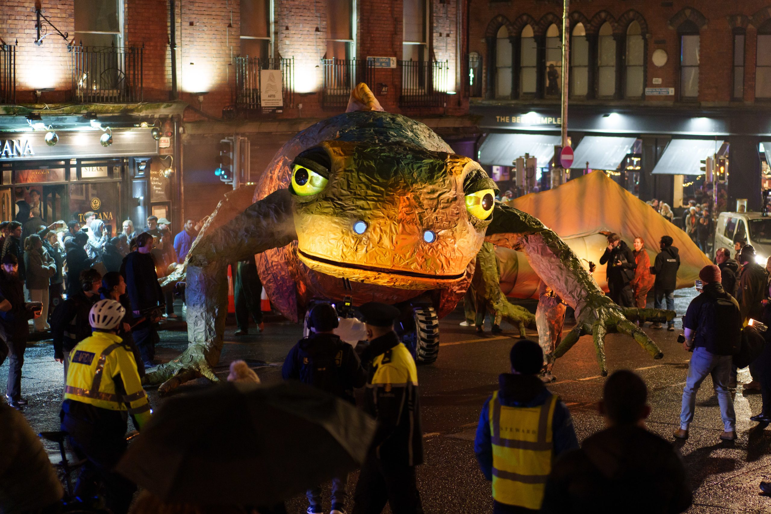 Large newt puppet walking on street at night surrounded by onlookers