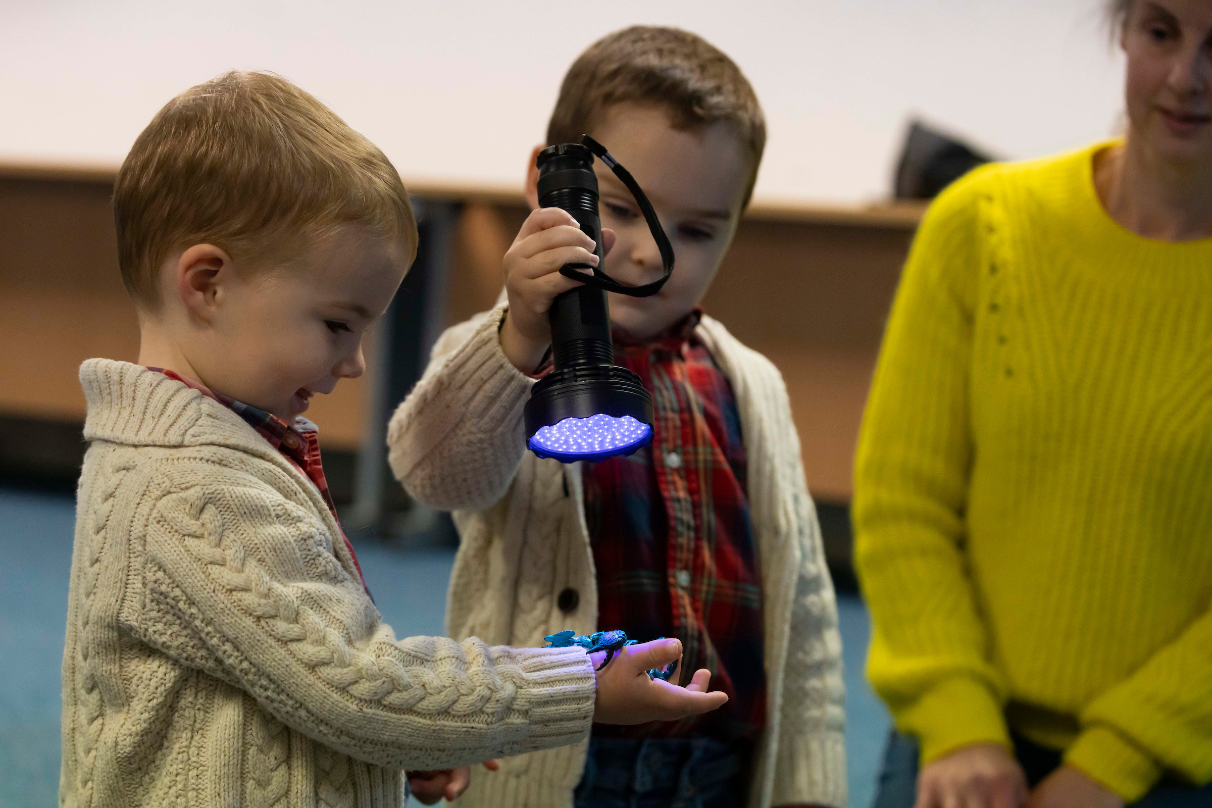 Two students using a UV flashlight to illuminate an object on one child’s hand, with an adult in a bright yellow sweater nearby.