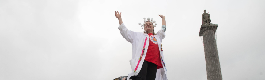 Person wearing a lab coat performing on jumping stilts in a town square with a tall monument in the background