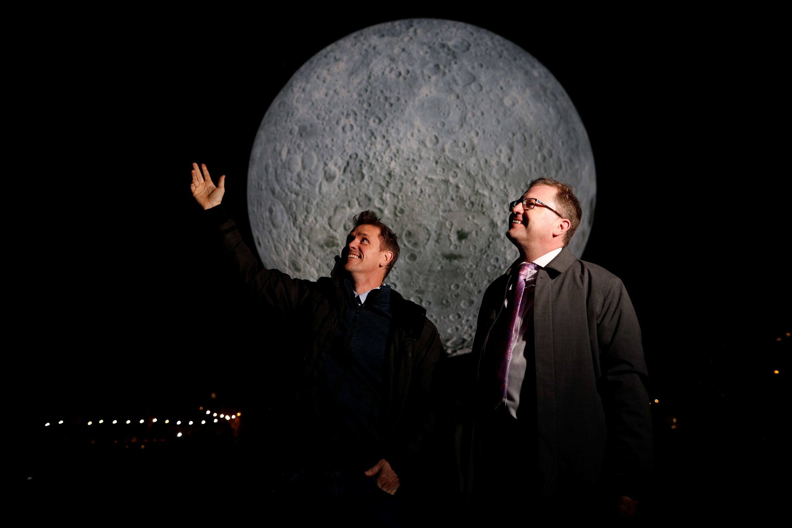 Two people stand outdoors at night pointing toward a large illuminated moon installation.