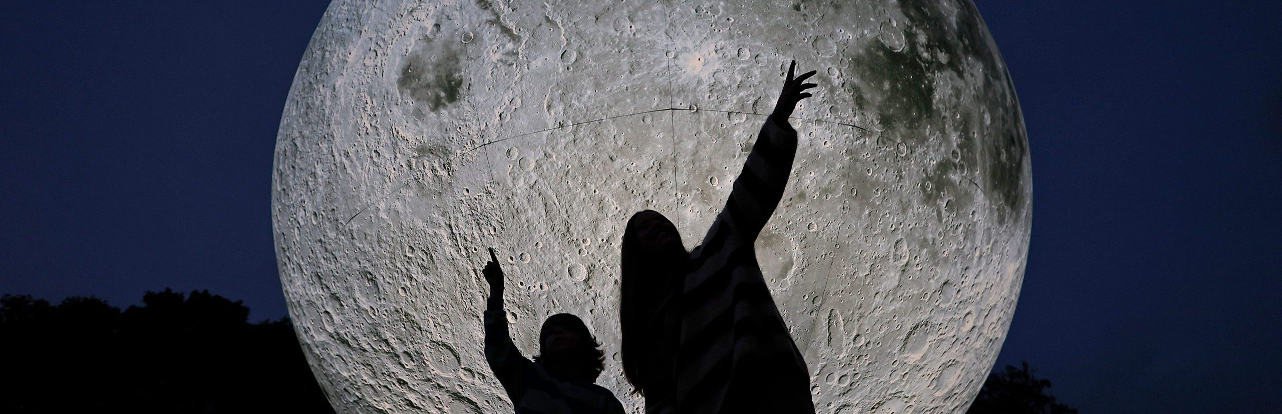 Silhouetted figures reach up toward a large illuminated moon installation at night.