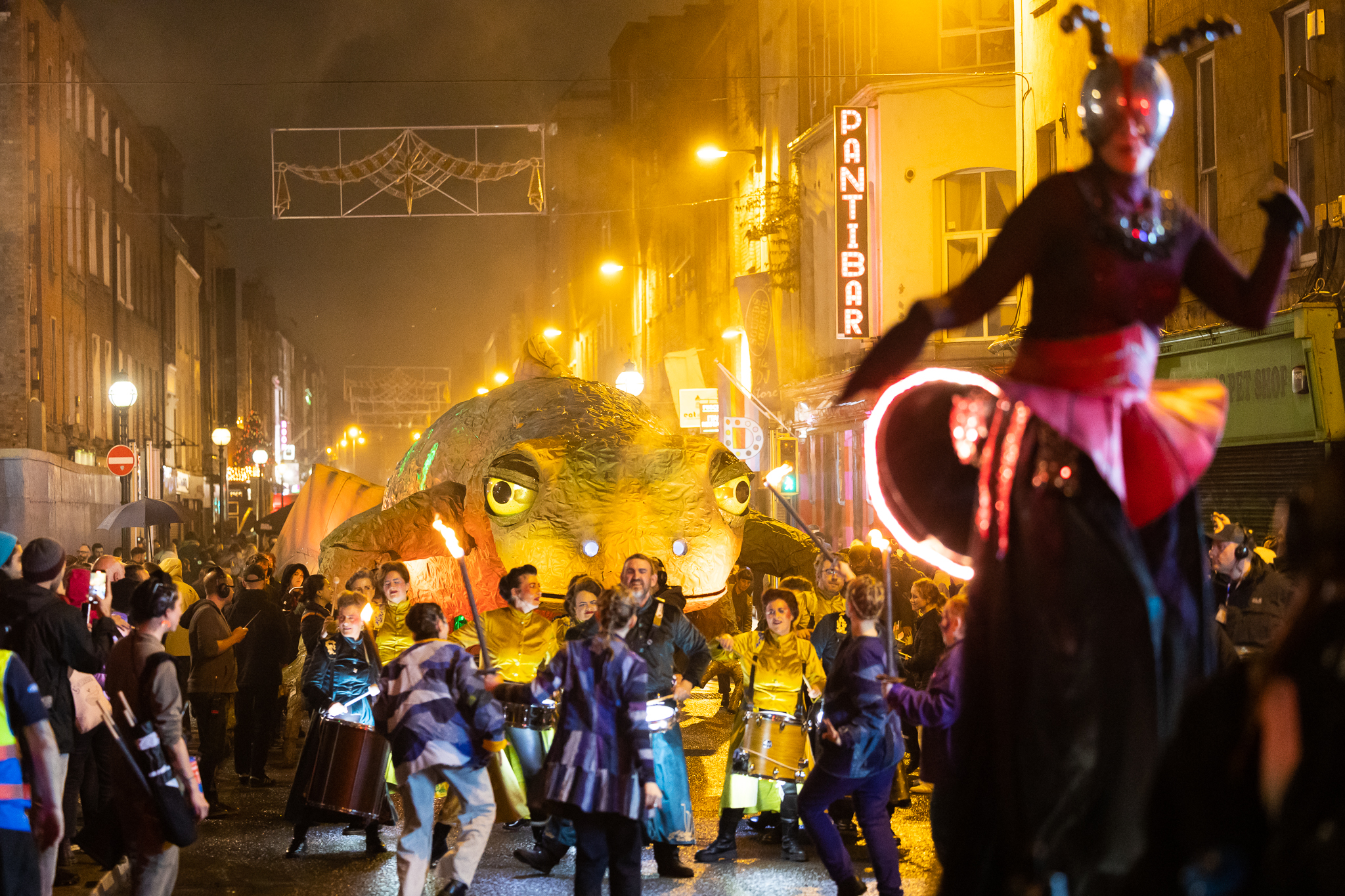Night-time street parade with drummers and a large illuminated creature, surrounded by crowds under glowing streetlights.